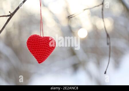 Valentine Herz im Winterwald gegen die Sonne. Rot gestricktes Herz hängt an einem Ast, Symbol der romantischen Liebe, Hintergrund für den Winterurlaub Stockfoto