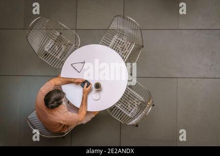 Mann sitzt am runden weißen Tisch auf Smartphone mit Kaffee von oben gesehen Stockfoto