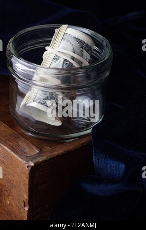 Eine Rolle von 100 us-Dollar-Banknoten in Glas auf Holzschublade mit blauem Samt auf dunklem launischem schwarzem Hintergrund, Nahaufnahme, Kopierraum, Finanzierung und Stockfoto