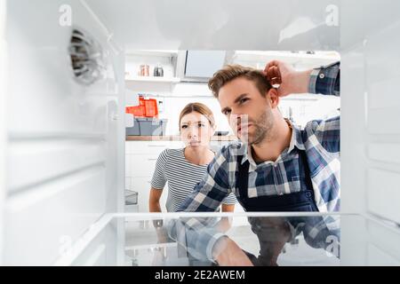 Nachdenklicher Handwerker und junge Frau, die auf dem Gefrierschrank auf verschwommen schauen Vordergrund in der Küche Stockfoto