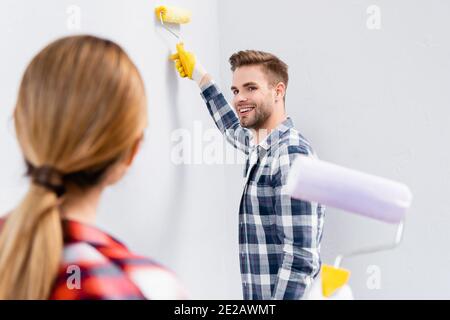 Glücklicher junger Mann mit Walzenbemalung Wand beim Blick auf Verschwommene Frau im Vordergrund drinnen Stockfoto