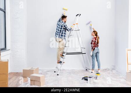 Voller Länge lächelnd junges Paar mit Rollen Malerei Wand Und einander in der Nähe der Leiter zu Hause angeschaut Stockfoto