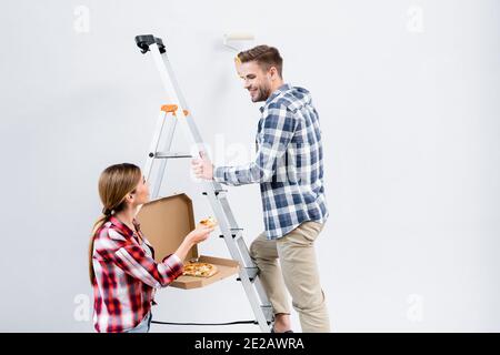 Lächelnder junger Mann mit Walzenmalerei Wand auf Frau Geben Stück Pizza neben Leiter isoliert auf grau Stockfoto