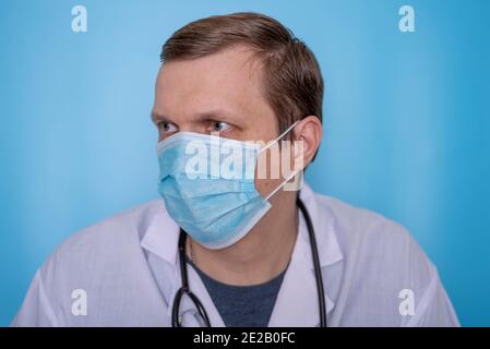 Ein männlicher Virologe in Maske und Morgenmantel auf blauem Hintergrund. Das Konzept von Gesundheit und Behandlung. Stockfoto