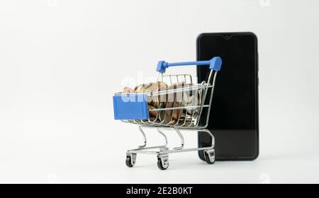 Warenkorb voller Münzen mit einem Smartphone.Geld und Finanzkonzept, E-Commerce, Business-Marketing, Shopping, Mini-Supermarkt-Trolley auf Isolat Stockfoto