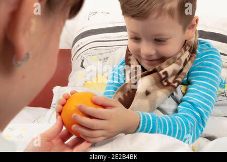 Mama gibt einem kranken Kind vorsichtig einen Jungen eingehüllt Ein warmer Schal mit einer frischen Orange Stockfoto