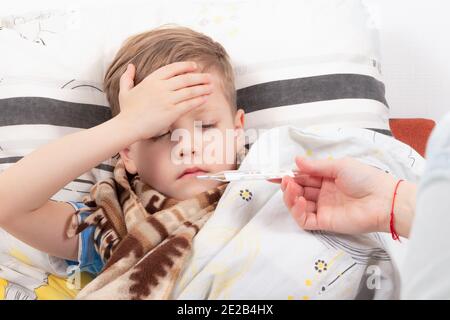 Ein kleiner Junge, ein Kind in einen Schal gehüllt, ist zu Hause krank, liegt im Bett und hält sich an seine Stirn, Mama gibt ihm ein Thermometer Stockfoto