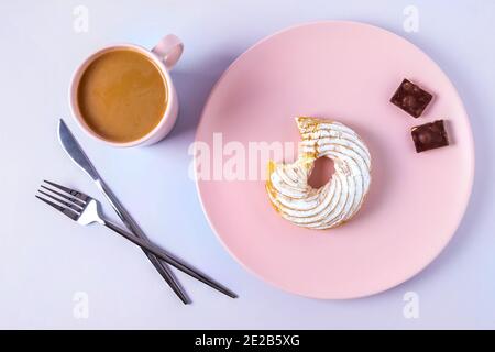 Draufsicht auf Stillleben eines gebissenen Kuchens auf einem rosafarbenen Teller, Besteck und einer Tasse Kakao oder Kaffee mit Milch. Selektiver Fokus, horizontale Ausrichtung. Stockfoto