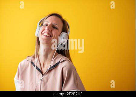 Fröhliche junge Frau, die freudig lacht, während sie mit ihren Kopfhörern auf dem Kopf Musik hört. Auf gelbem Hintergrund. Stockfoto