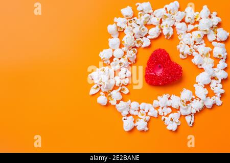 Popcorn und eine Kerze in Form eines roten Herzens auf orangefarbenem Hintergrund mit leerem Raum. Stockfoto