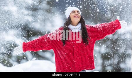 Mit weit geöffneten Armen genießt Frau in rotem Pullover, weißen Handschuhen, Schal und Hut an einem verschneiten Wintertag frische Luft. Stockfoto