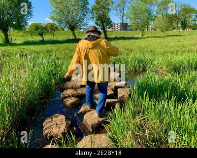 Ein Mann mit braunem Hut, gelbem Regenmantel und blauer Jeans tritt vorsichtig auf Trittsteine über ein kleines Wasser. Holländische Landschaft mit Deich. Stockfoto