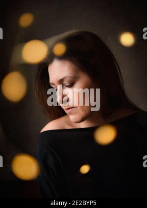Nachdenkliche hübsche Frau in dunklem Zimmer mit hellem Bokeh Umgebung Sie Stockfoto