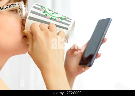 Frau trinkt aus einer Tasse und schaut auf ihr Handy. Stockfoto