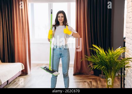 In voller Länge Foto von positiven fröhlich Hausfrau tragen gelbe Gummihandschuhe, genießen Waschboden halten Mopp fühlen sich zufrieden tragen Denim Jeans casual cl Stockfoto