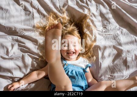 Fröhliches Baby lachend.EIN fröhliches Mädchen liegt auf dem Bett und schreit mit Freude/Bewegung und Fitness auf der Tagesdecke liegend. Stockfoto