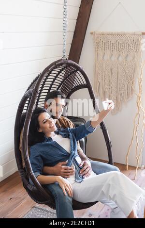 Glücklich junges Paar sitzt in hängen Rattan Stuhl und nehmen Selfie auf dem Handy Stockfoto