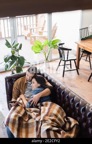 Blick in den hohen Winkel des jungen Paares beim Fernsehen auf der Ledercouch unter der karierten Decke, verschwommener Vordergrund Stockfoto