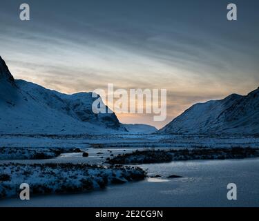 Blick nach Norden durch den Pass von Glencoe bei Sonnenuntergang an einem verschneiten Wintertag. Rotwild ernähren sich in der Ferne unterhalb der drei Schwestern von Glencoe. Stockfoto