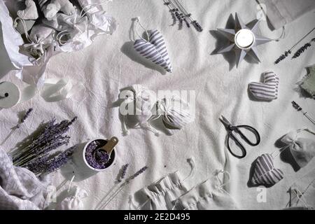 Handgemachte Lavendelsäckchen in Textiltaschen und weichen Herzen. Trockene Lavendelblüten zum Füllen, Scheren und Kerzen. Flacher Lay auf Leinentextilien Stockfoto