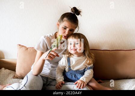 Lustige Kinder spielen mit einem Papagei. Zwei Mädchen und ein Teenager zu Hause auf dem Bett, das Kind kümmert sich um die Haustiere Stockfoto