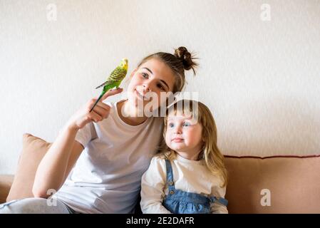 Lustige Kinder spielen mit einem Papagei. Zwei Mädchen und ein Teenager zu Hause auf dem Bett, das Kind kümmert sich um die Haustiere Stockfoto