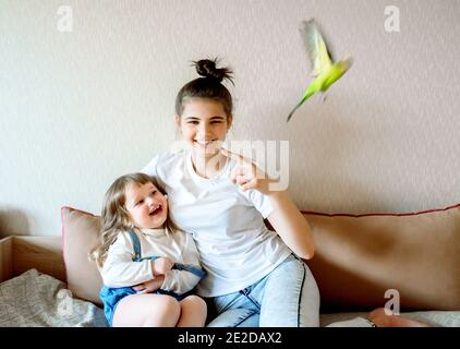 Lustige Kinder spielen mit einem Papagei. Zwei Mädchen und ein Teenager zu Hause auf dem Bett, das Kind kümmert sich um die Haustiere Stockfoto