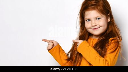 Lächelnd kleine rothaarige Mädchen mit Sommersprossen in gelb bequem longsleeve stehend und mit den Fingern zur Seite zeigend Stockfoto