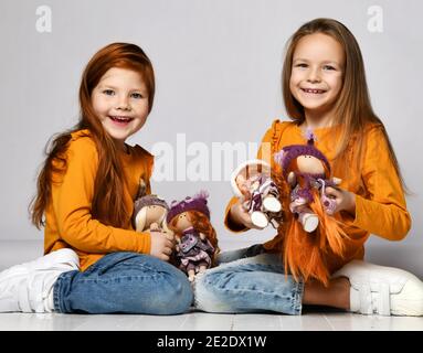 Lächelnde kleine Mädchen Schwestern oder Freunde in gelben Longsleeves, Jeans und Sneakers sitzen auf dem Boden und spielen mit Spielzeugpuppen Stockfoto