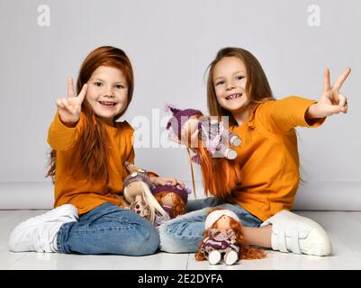 Lächelnde kleine Mädchen Schwestern in gelben Longsleeves, Jeans und Sneakers sitzen auf dem Boden und spielen mit Puppen und zeigen Friedenszeichen Stockfoto