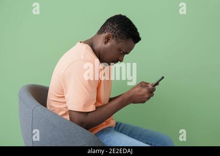 Ein afroamerikanischer Mann mit einem Buckel und einem Mobiltelefon, das auf einem Stuhl in der Nähe sitzt Farbwand Stockfoto