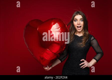 Überrascht attraktive junge Mädchen mit Vorlage Herz geformten Ballon auf rotem Hintergrund, kopieren Raum für valentinstag Anzeige Stockfoto