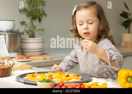 Nettes kleines Mädchen 2-4 in grauem Kleid Kochen Pizza in der Küche, ordnet Kind Zutaten auf Pizza Basis Stockfoto