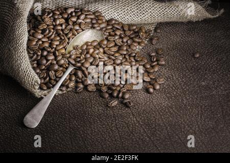 Geröstete Kaffeebohnen schokoladig getöntes Stillleben mit einer hessischen Tasche und einem Vintage-Metalllöffel auf Büffelleder. Lager Stockfoto