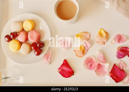 Frühstück im Bett mit Makronen. Tasse duftenden Kaffee, Untertasse mit Dessert und Kirschen auf dem Tisch. Rosenblüten auf weißem Tablett Überraschung zum Valentinstag. Draufsicht. Stockfoto