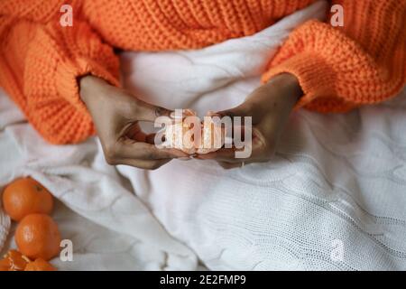 Afro Frau Hände schälen reife süße Mandarine, tragen orange Pullover, liegen im Bett unter dem Plaid Stockfoto