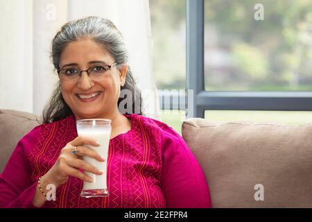 EINE GLÜCKLICHE ALTE FRAU, DIE AUF DEM SOFA SITZT UND MILCH TRINKT Stockfoto