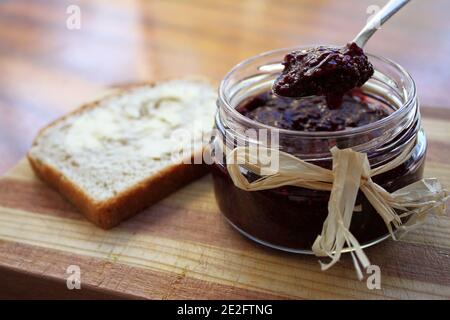 Ein Löffel Marmelade aus einem Glas neben einem Scheibe frisches Brot auf einem Holzbrett Stockfoto