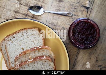 Eine Marmelade aus Glas neben einem Teller Brot Und einen Löffel vor einer rustikalen Holzkulisse Stockfoto