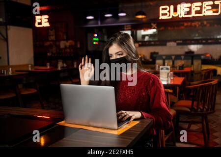 Porträt einer jungen Frau, die während eines Videoanrufs mit der Hand auf ihren Laptop schwenkt, während sie in einem Restaurant sitzt und eine Covid 19 Schutzmaske trägt. Stockfoto