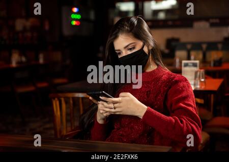 Porträt einer Frau, die in einem Restaurant mit Covid 19 Schutzmaske sitzt und ihr Telefon benutzt. Stockfoto