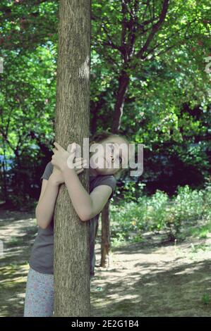 Blonde Kind, Mädchen, 8 Jahre alt, Blick nach oben, hinter einem Baum stehen und umarmt es im Wald, Vorderansicht, selektive Fokus Stockfoto