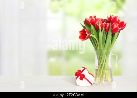 Alles Liebe zum Valentinstag oder Muttertag Hintergrund. Schöne rote Tulpen und eine Geschenkbox in Form eines Herzens auf dem Tisch über verschwommenem natürlichen Hintergrund. Platz für Stockfoto