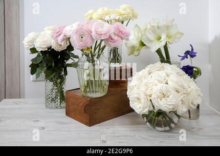 Weiße Rosen, Ranunculus, blaue Anemonen, gelbliche Butterblumen, Lilien in runden Vasen auf den Holzkisten auf dem Tisch für einen besonderen Anlass als Kitch Stockfoto