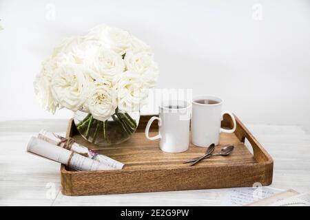 Ein Strauß weißer Rosen in einer runden Glasvase auf einem Holztablett ein Tisch mit zwei Tassen Tee und einem eingewickelten Papierrollen. Speicherplatz kopieren Stockfoto