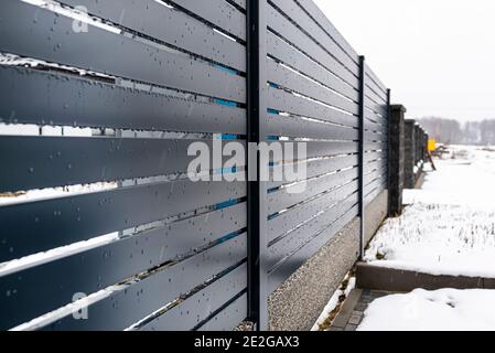 Moderne anthrazitfarbene Einzäunung, mit sichtbarem Zaunfundament-Anschluss, es regnet im Winter, im Hintergrund liegt Schnee auf dem Boden. Stockfoto