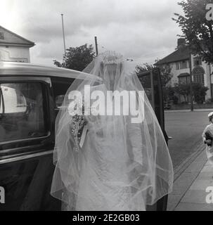 1960s, historisch, eine Braut in ihrem Hochzeitskleid und Kopfstück mit langem Schleier bedeckt ihr Gesicht, tritt aus ihrer Hochzeit oder Brautwagen, Ankunft für die cermony, England, Großbritannien. Der lang bedeckende Gesichtsschleier symbolisierte traditionell Bescheidenheit und Gehorsam und war eine elegante und allgemein getragene Brautkleider in dieser Zeit. Stockfoto