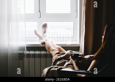 Junge Frau, die sich auf einem Sessel neben dem Fenster ausruhte und heißen Tee in einer Decke trank. Hochwertige Fotos Stockfoto