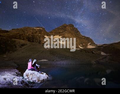 Landschaft von Bergen mit Schnee bei Nacht in den Alpen. Wunderschöner Bergrücken mit hohen felsigen Gipfeln, Paar sitzt auf Stein in der Nähe des Sees, genießen Milchstraße mit leuchtenden Sternen am Himmel, Matterhorn. Stockfoto