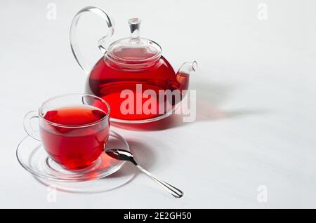 Roter Hibiskustee in transparenter Teekanne mit Tasse im Sonnenlicht mit Schatten auf weißem Holzbrett, Kopierraum. Stockfoto